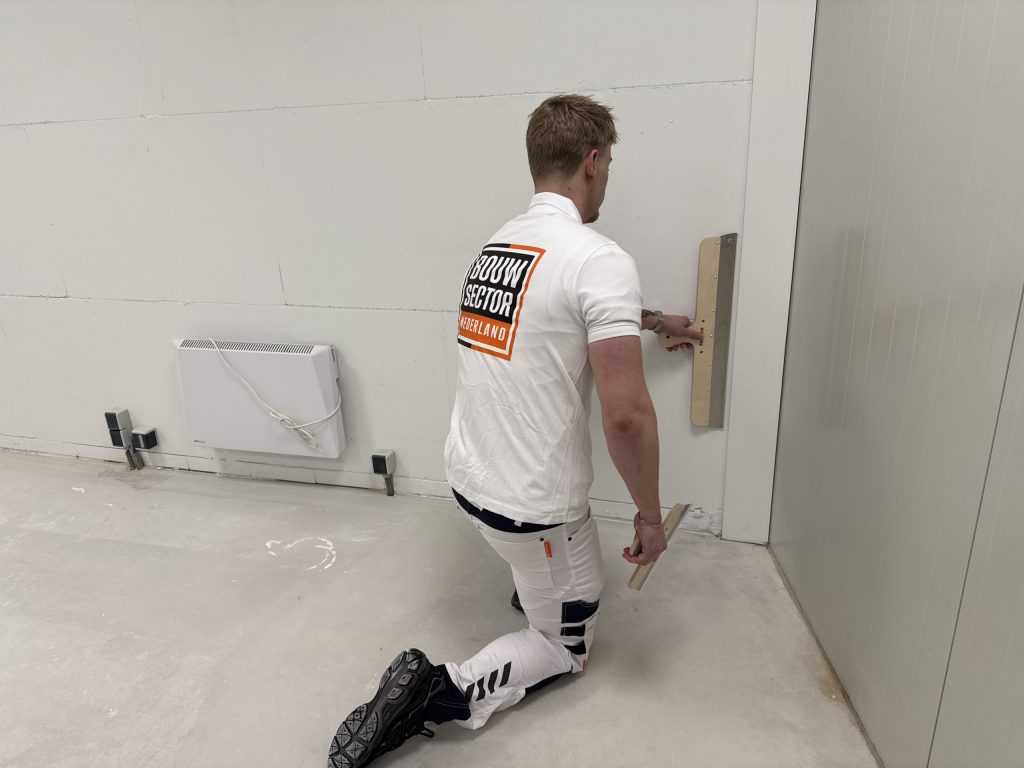 Stukadoor van Bouwsector Nederland werkt geknield de onderkant van een wand bij met een brede spaan