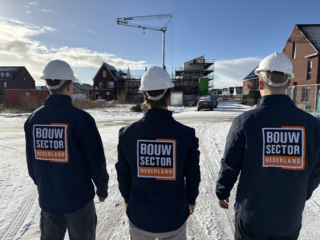 Drie vakmensen van Bouwsector Nederland in werkjassen met logo en witte helm op een besneeuwde bouwplaats
