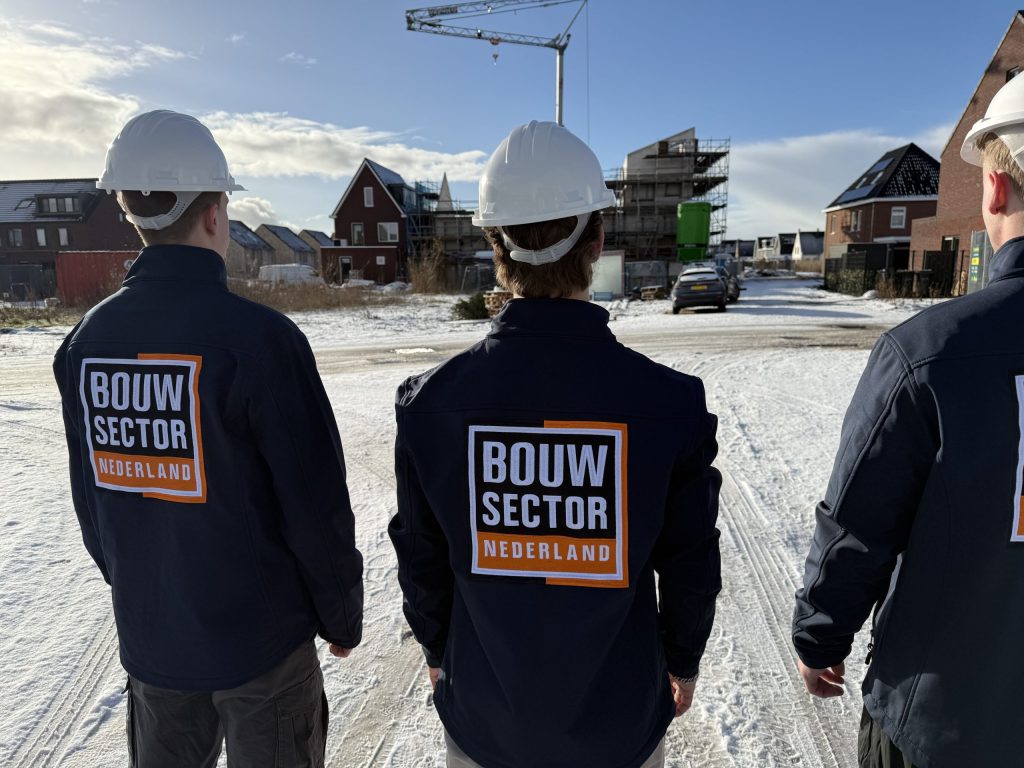 Drie vakmensen van Bouwsector Nederland met witte helm op een besneeuwde straat met woningen in aanbouw